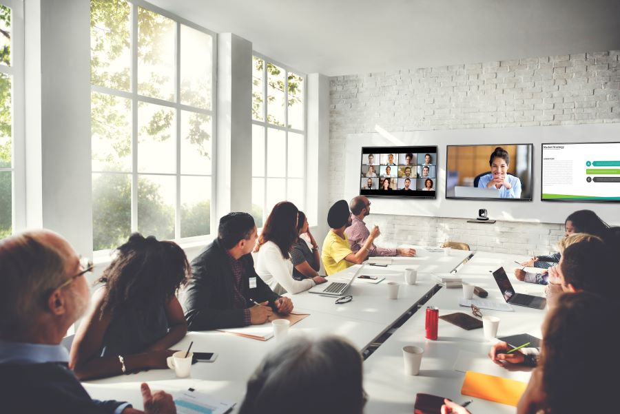 A large meeting room filled with employees looking at a remote meeting display on the TV at the front of the room.