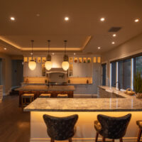 A modern kitchen featuring a large island with bar stools, recessed and pendant lighting, and large windows showing outdoor greenery.