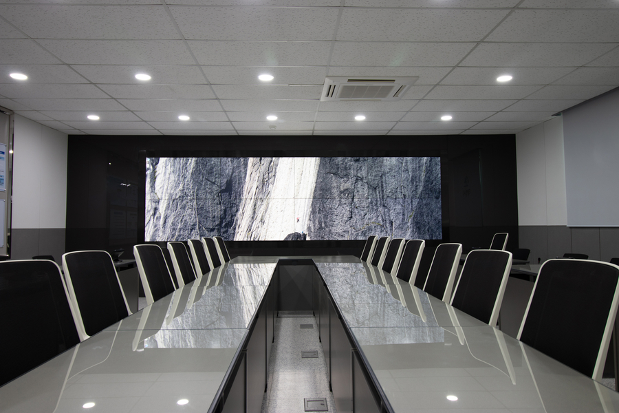 Modern conference room with a sleek glass table, ergonomic black chairs, and a large video wall displaying a high-resolution image of a rock climber on a cliff face.