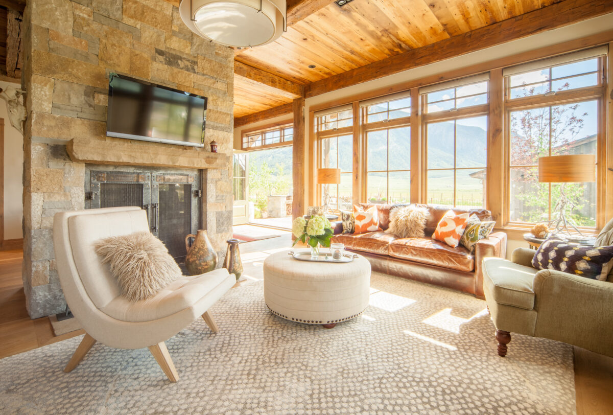 Bright, rustic living room with large windows, leather sofa, and mountain views, showcasing natural light streaming through uncovered windows.