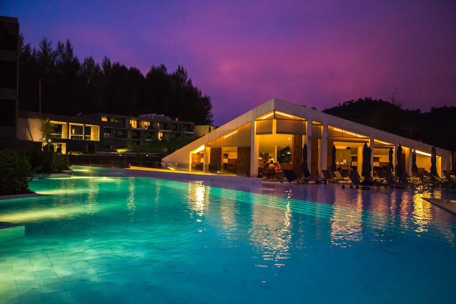 A large outdoor pool illuminated by pool lights. A large covered patio is next to the pool, illuminated by ceiling lights.