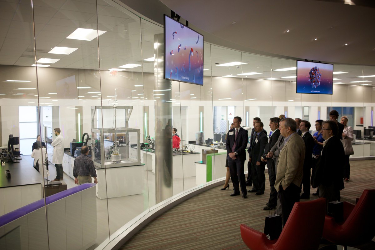 A group of professionally dressed visitors observes a modern laboratory through a curved glass wall, while two ceiling-mounted digital displays show colorful scientific graphics.