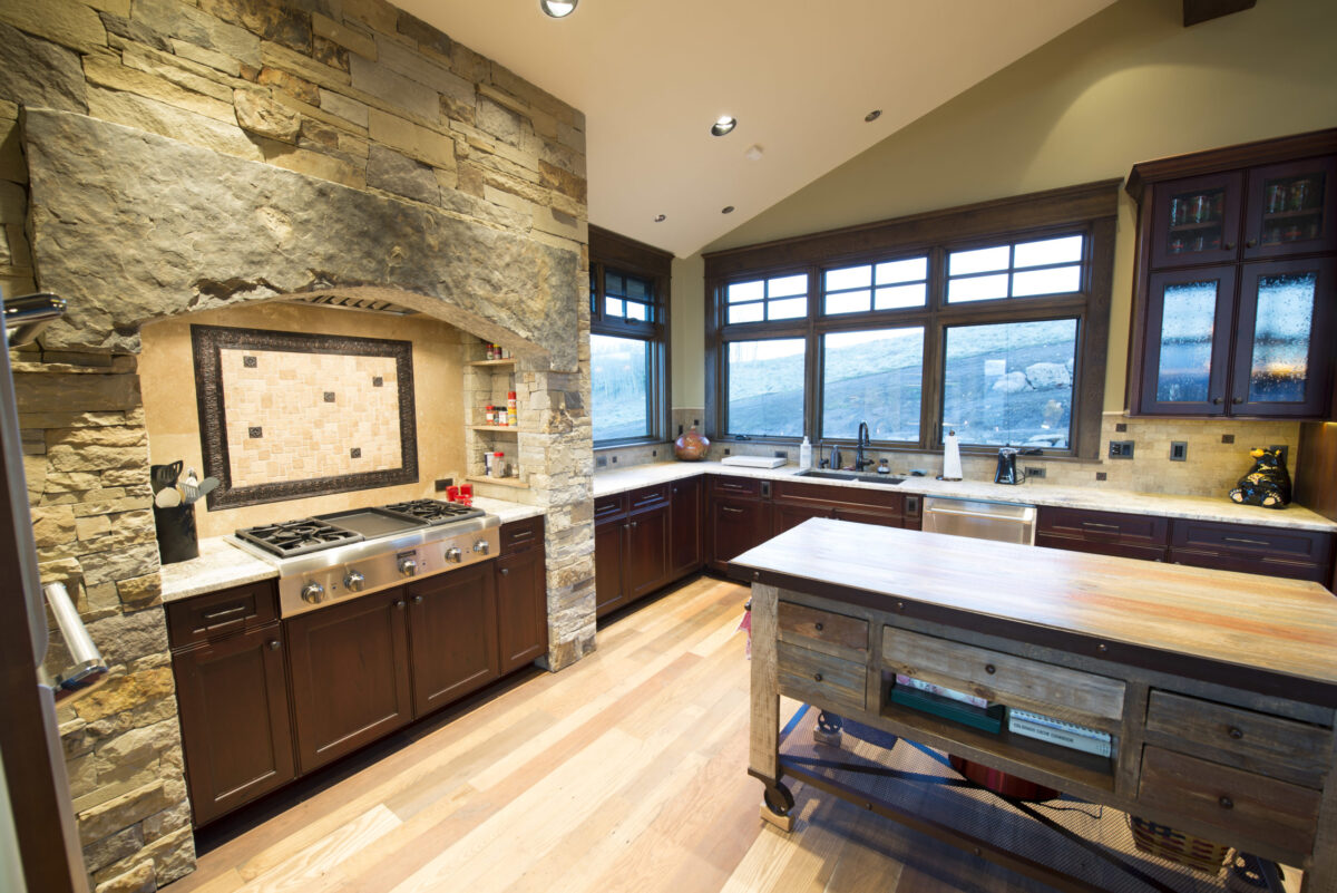 Rustic kitchen with stone accent wall, wooden cabinets, large windows, farmhouse island, and stainless steel gas stove.